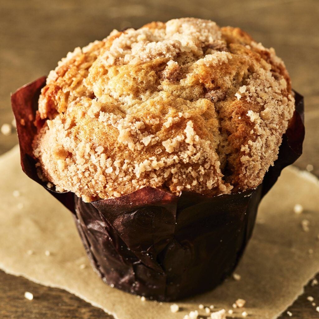 A golden-brown muffin with a crumbly sugar topping sits wrapped in dark brown paper on a parchment sheet, conveying a warm, freshly-baked feel.