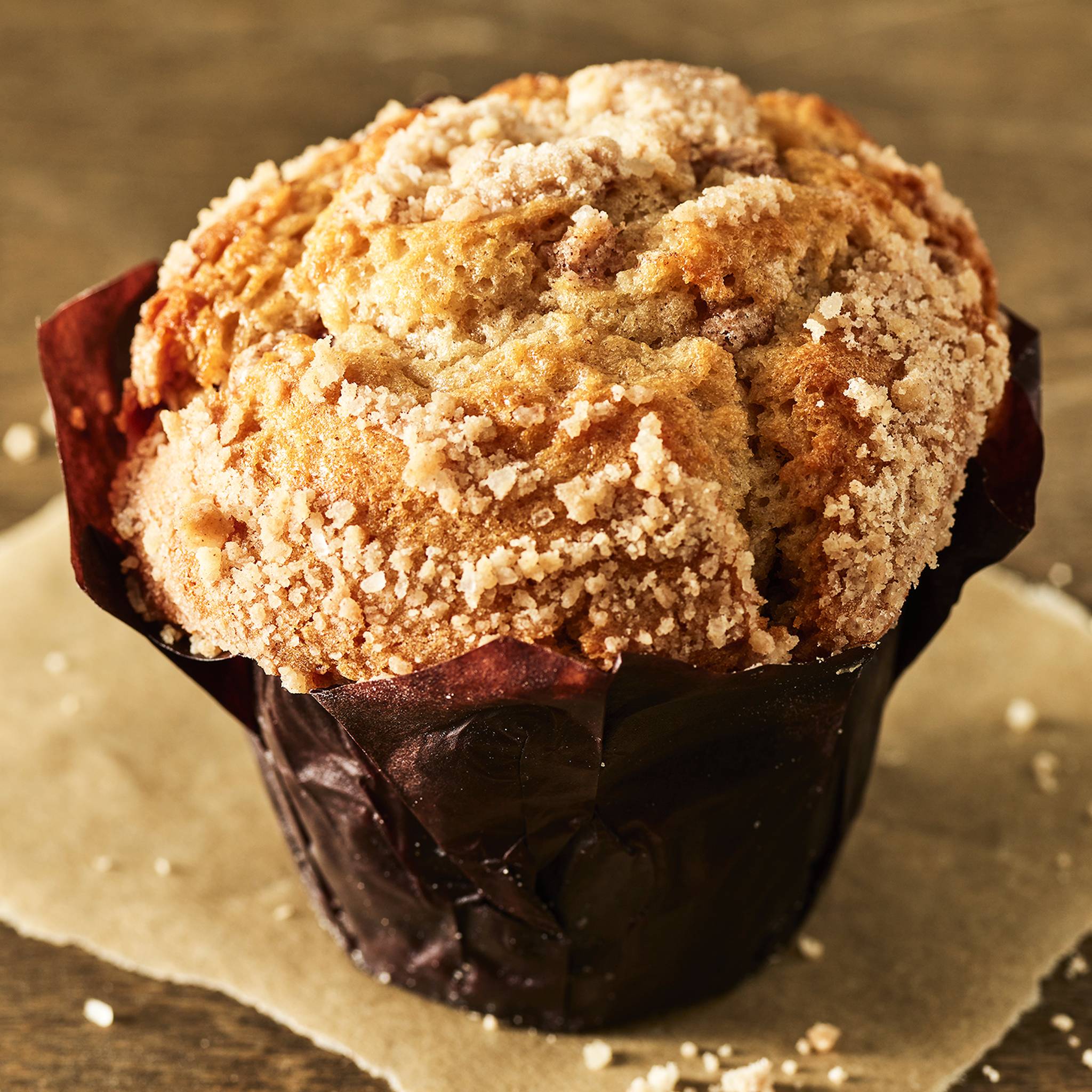 A golden-brown muffin with a crumbly sugar topping sits wrapped in dark brown paper on a parchment sheet, conveying a warm, freshly-baked feel.