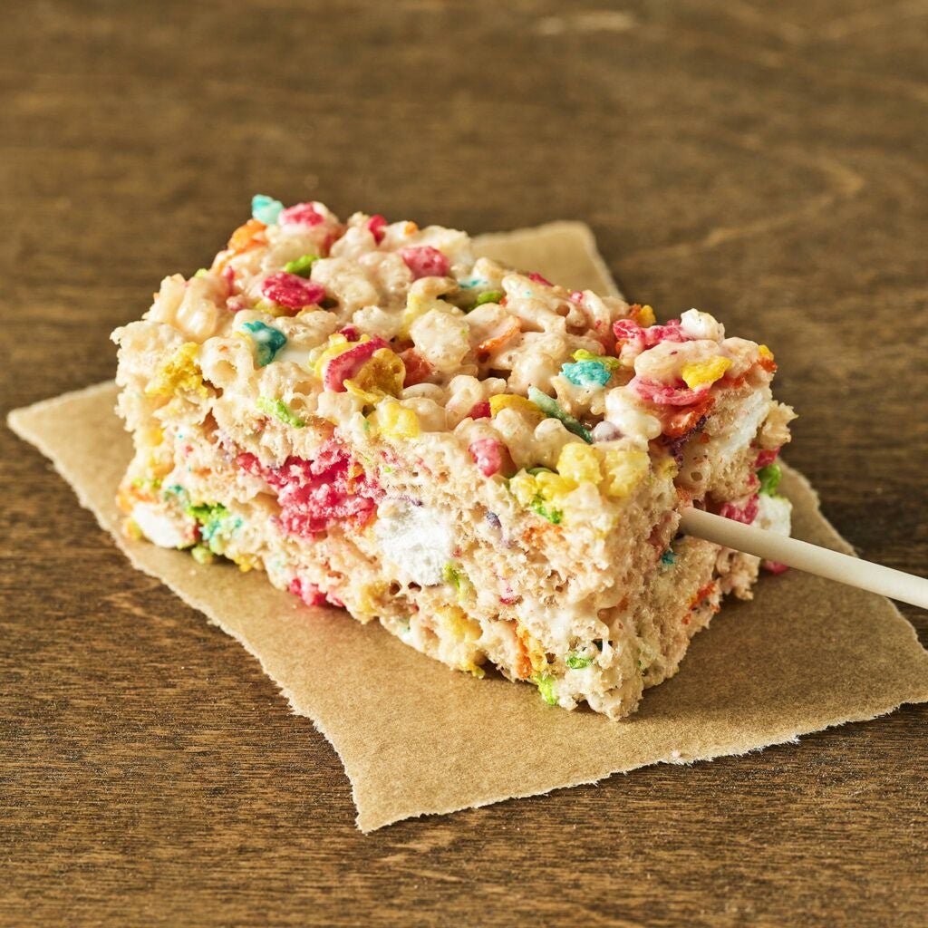 A colorful cereal treat bar with vibrant multicolored cereal pieces, held together by marshmallow, rests on brown parchment paper on a wooden surface.