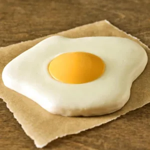 A realistic fried egg-shaped sugar cookie with a glossy yellow yolk center, rests on a brown paper, set against a wooden surface. The image is playful and whimsical.