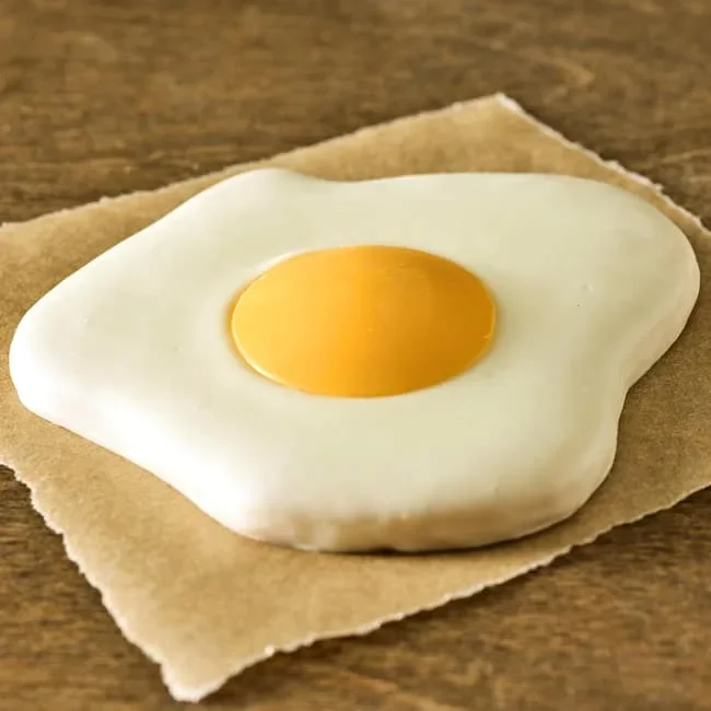 A realistic fried egg-shaped sugar cookie with a glossy yellow yolk center, rests on a brown paper, set against a wooden surface. The image is playful and whimsical.