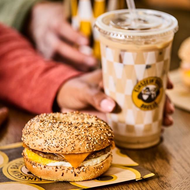 A hand holds an iced coffee in an Einstein Bagel Bros checkered cup. In the foreground, an everything bagel with sausage, egg, and cheese sits on branded packaging, evoking warmth.