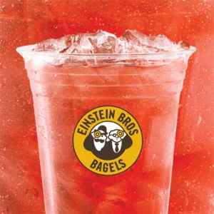 A clear plastic cup filled with iced red drink, showcasing the Einstein Bros Bagels logo. The vibrant background matches the drink's refreshing appearance.
